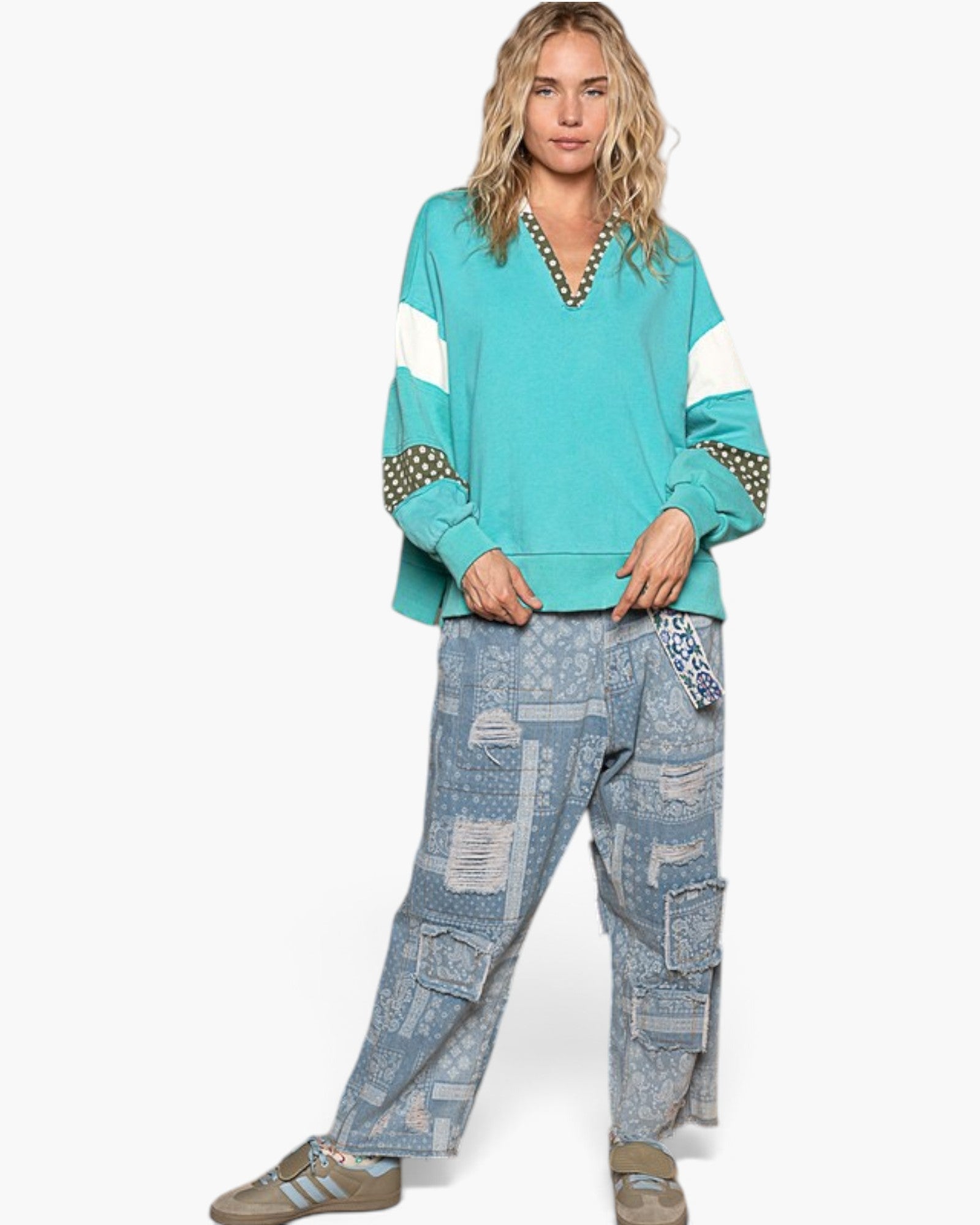A woman modeling a turquoise and white color-blocked knit sweater with patterned sleeve cuffs, paired with distressed light blue patchwork cargo pants and beige sneakers, against a plain white background.