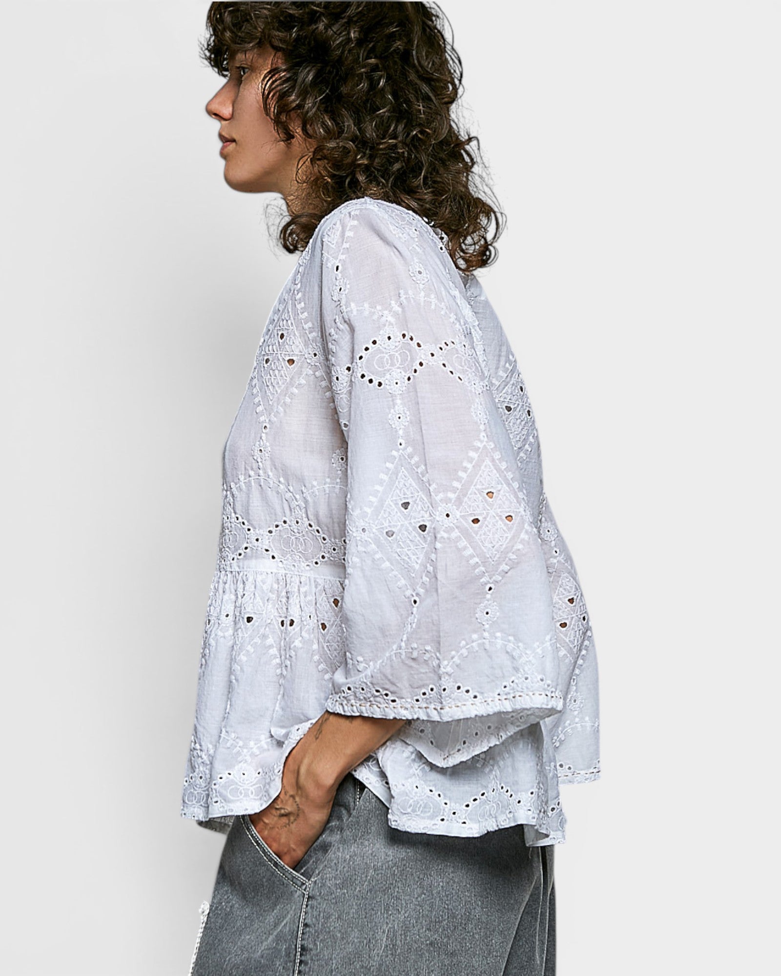 Woman wearing a white embroidered blouse and gray pants against a plain background