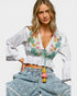 Woman wearing a white blouse with colorful embroidery and blue jeans with tassel details on a white background