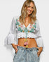 Woman wearing a white embroidered top with ruffles and denim shorts on a plain background