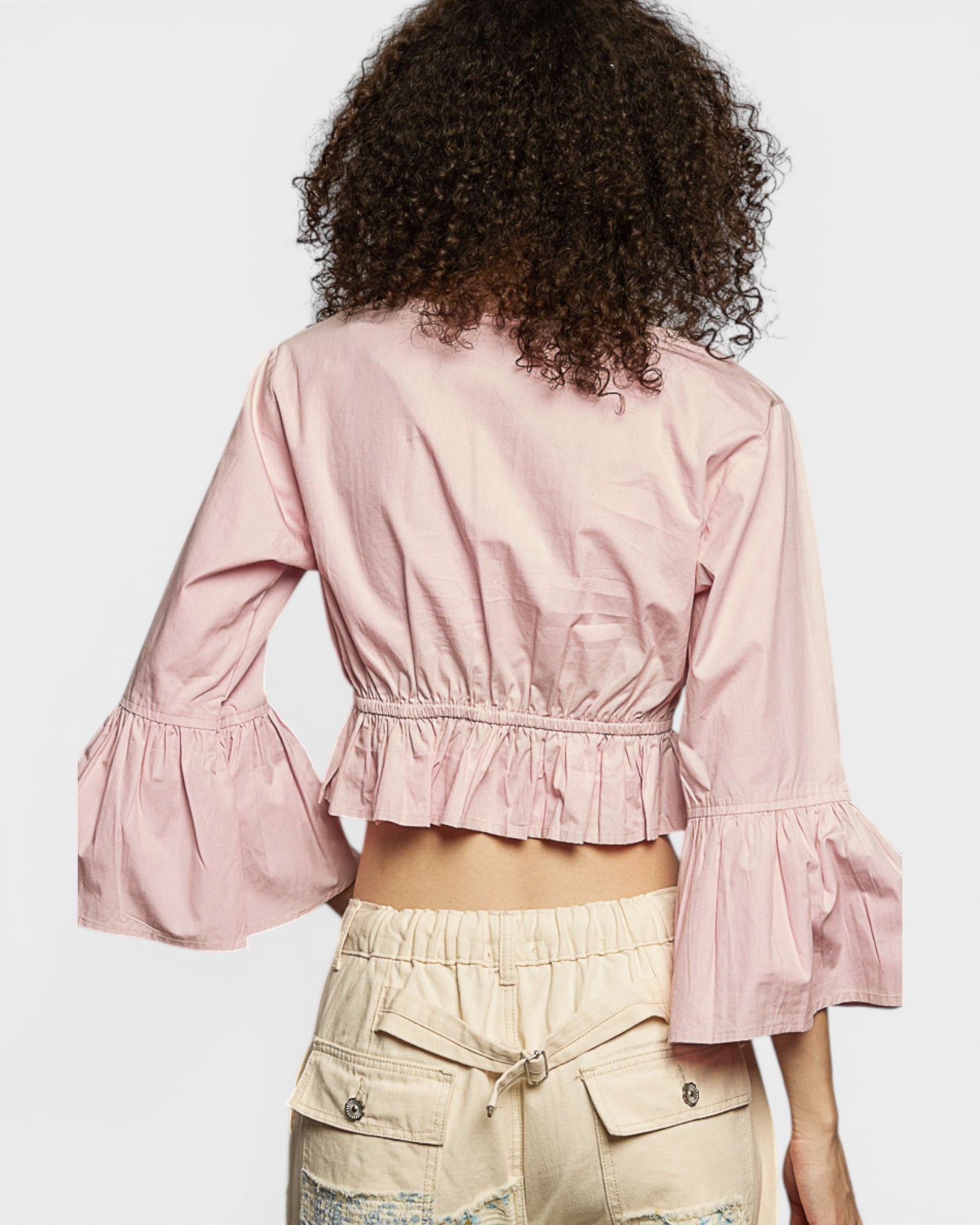 Person wearing a pink blouse with ruffled sleeves and beige pants on a light gray background