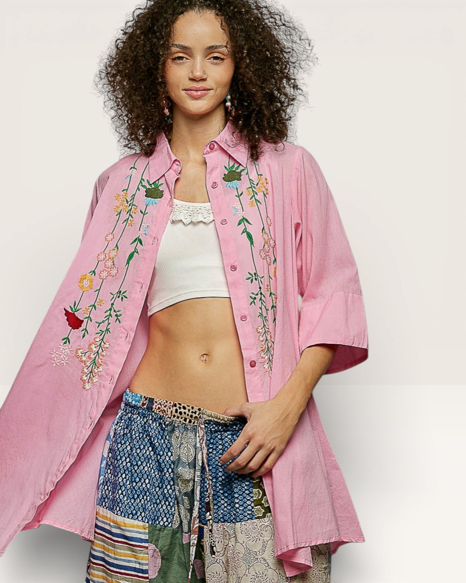 Woman wearing a pink embroidered shirt over a white top and patterned pants on a plain background