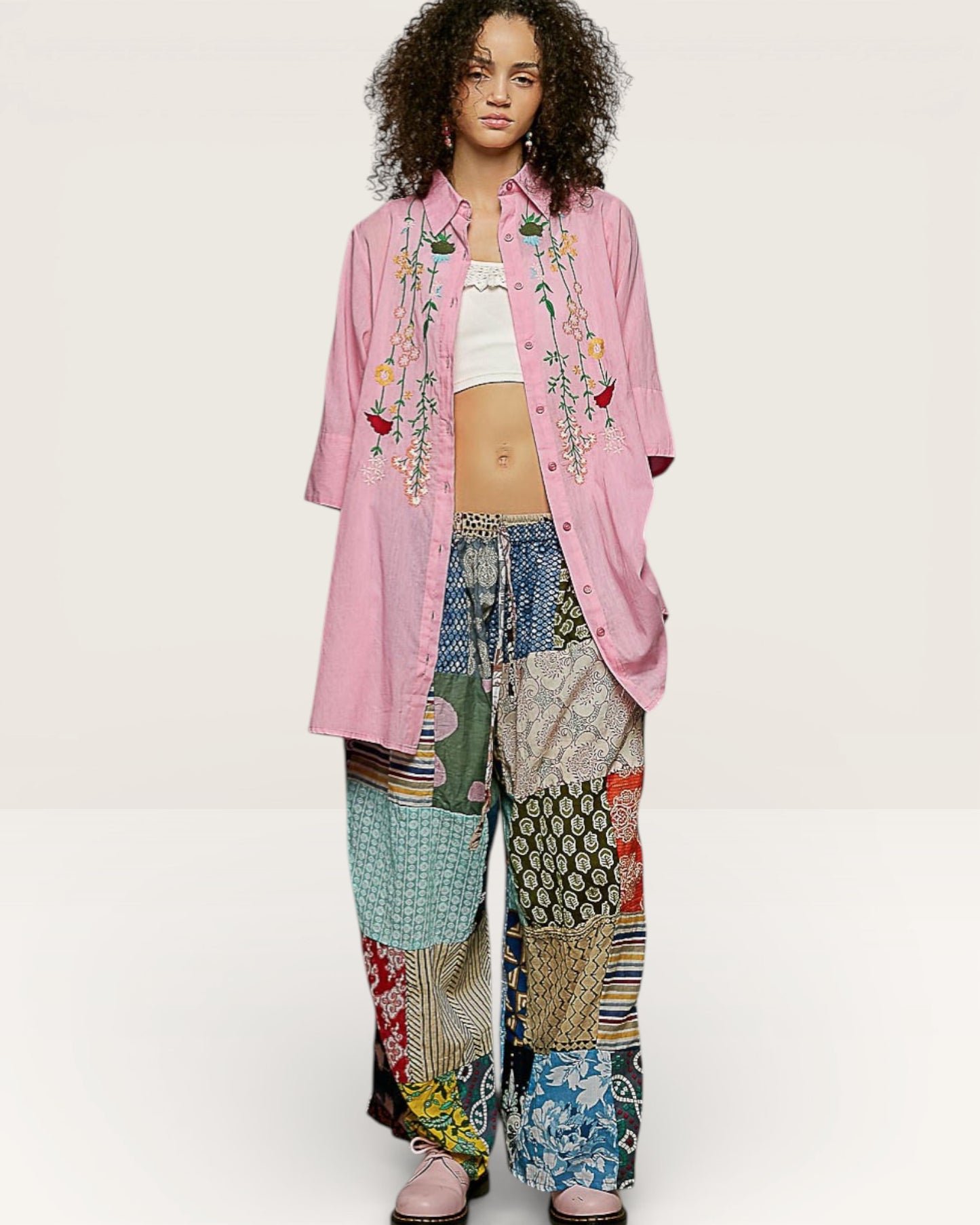 Person wearing a pink embroidered shirt and colorful patchwork pants on a white background