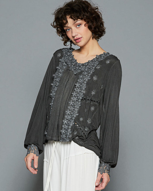 Woman wearing a dark gray embroidered blouse with white pants against a gray background