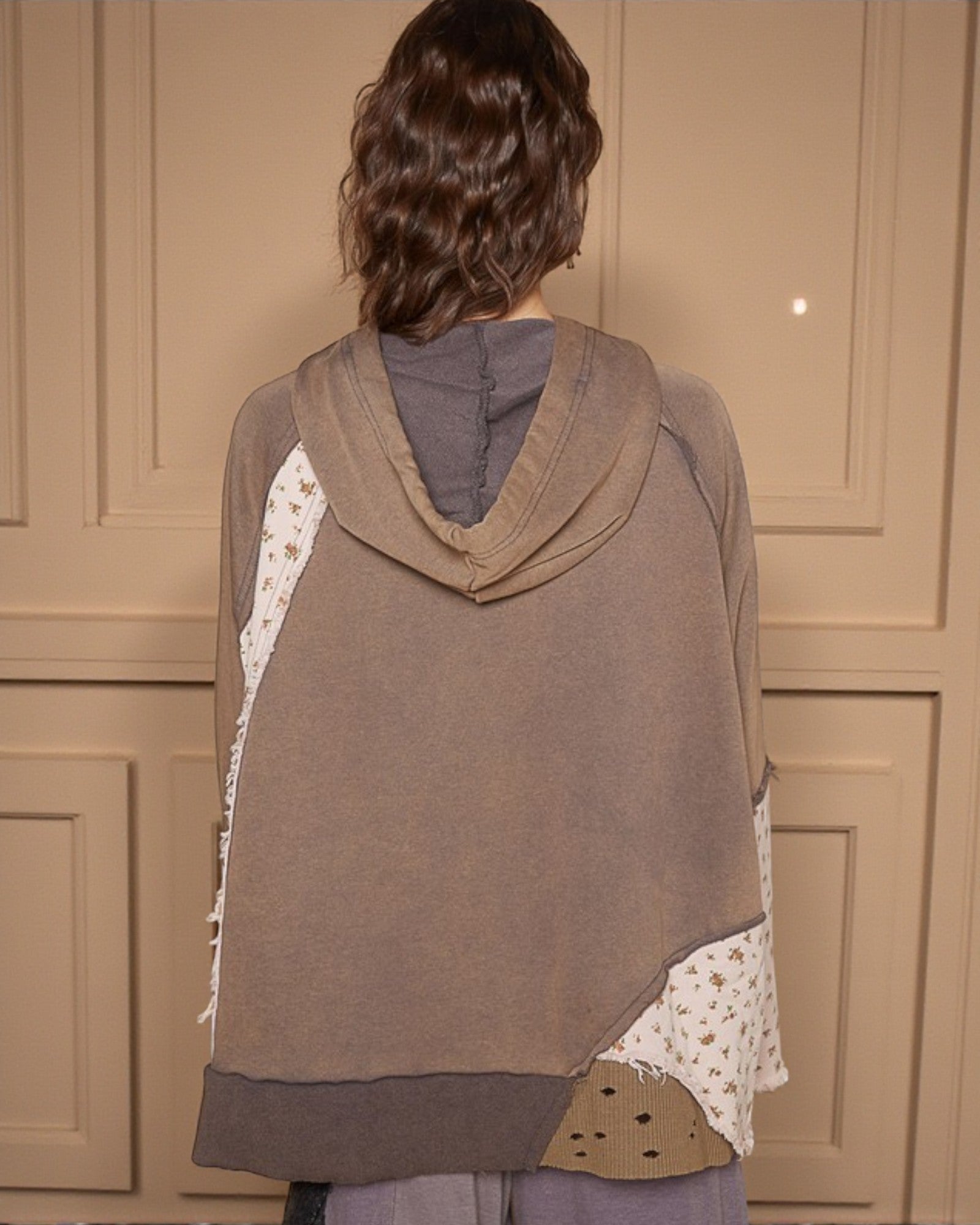 Person wearing a brown hooded garment with a patterned edge against a beige paneled wall.