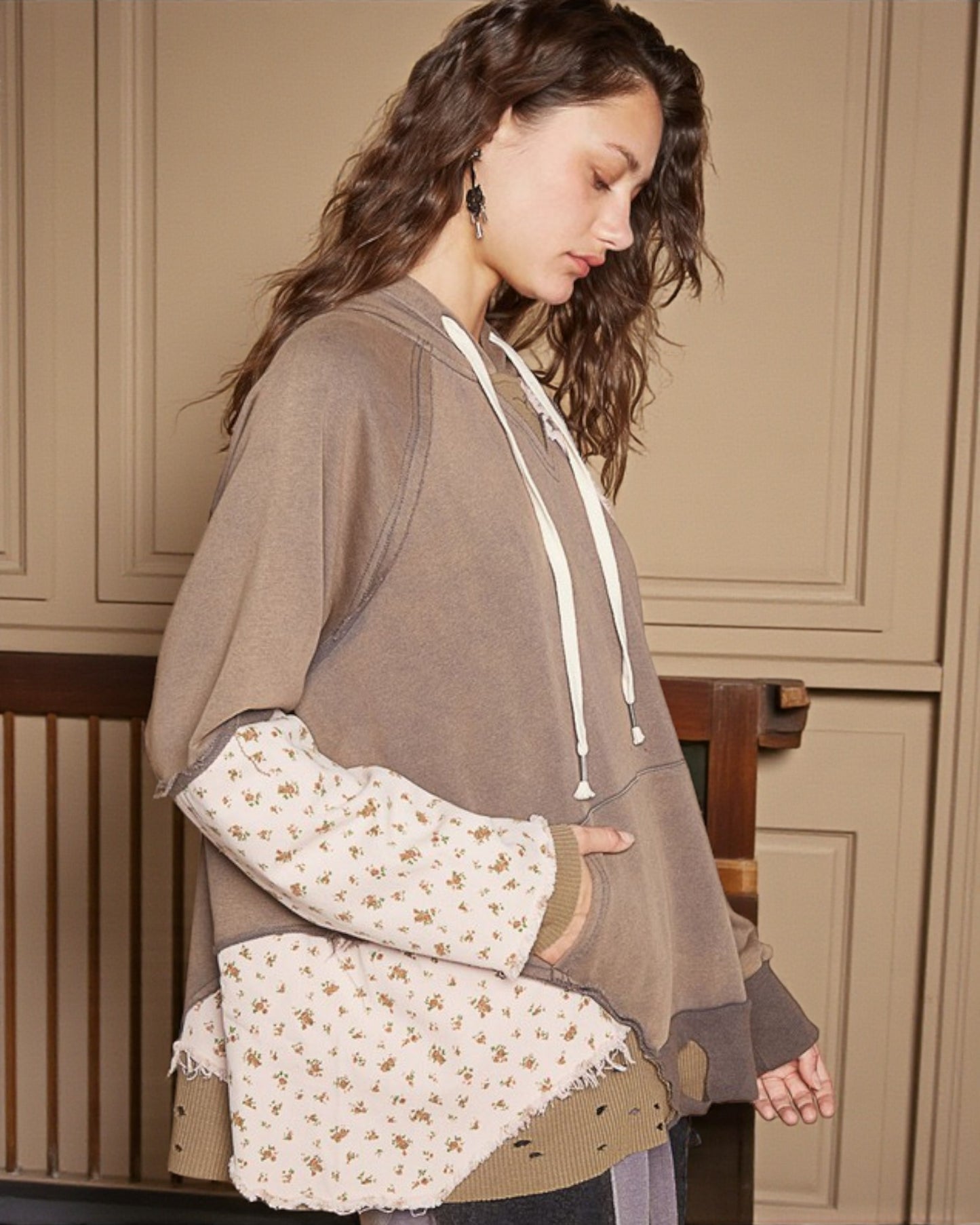 Woman wearing a brown hoodie with floral patterned sleeves against a beige wall.