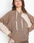 A woman wearing a brown pullover hoodie with cream-colored drawstrings and patterned knit sleeves, shown against a plain light background.