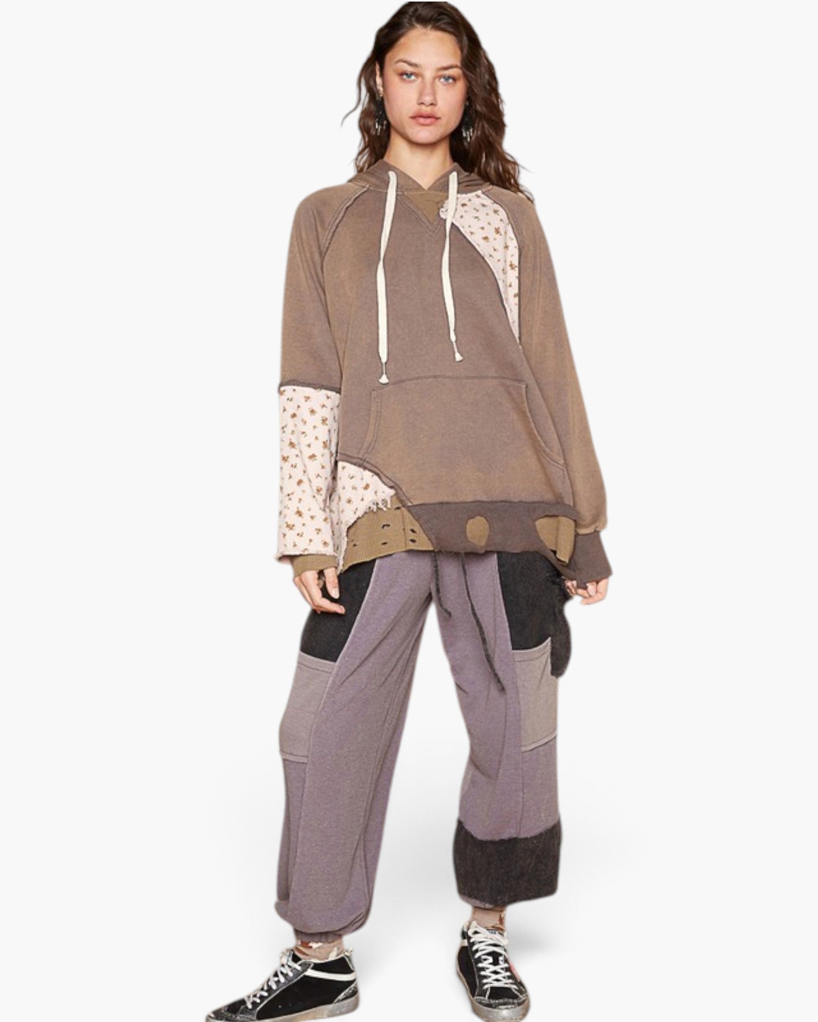 A model wearing a brown and beige patchwork hoodie with patterned sleeves, paired with matching wide-leg patchwork sweatpants and black sneakers, against a plain white background.