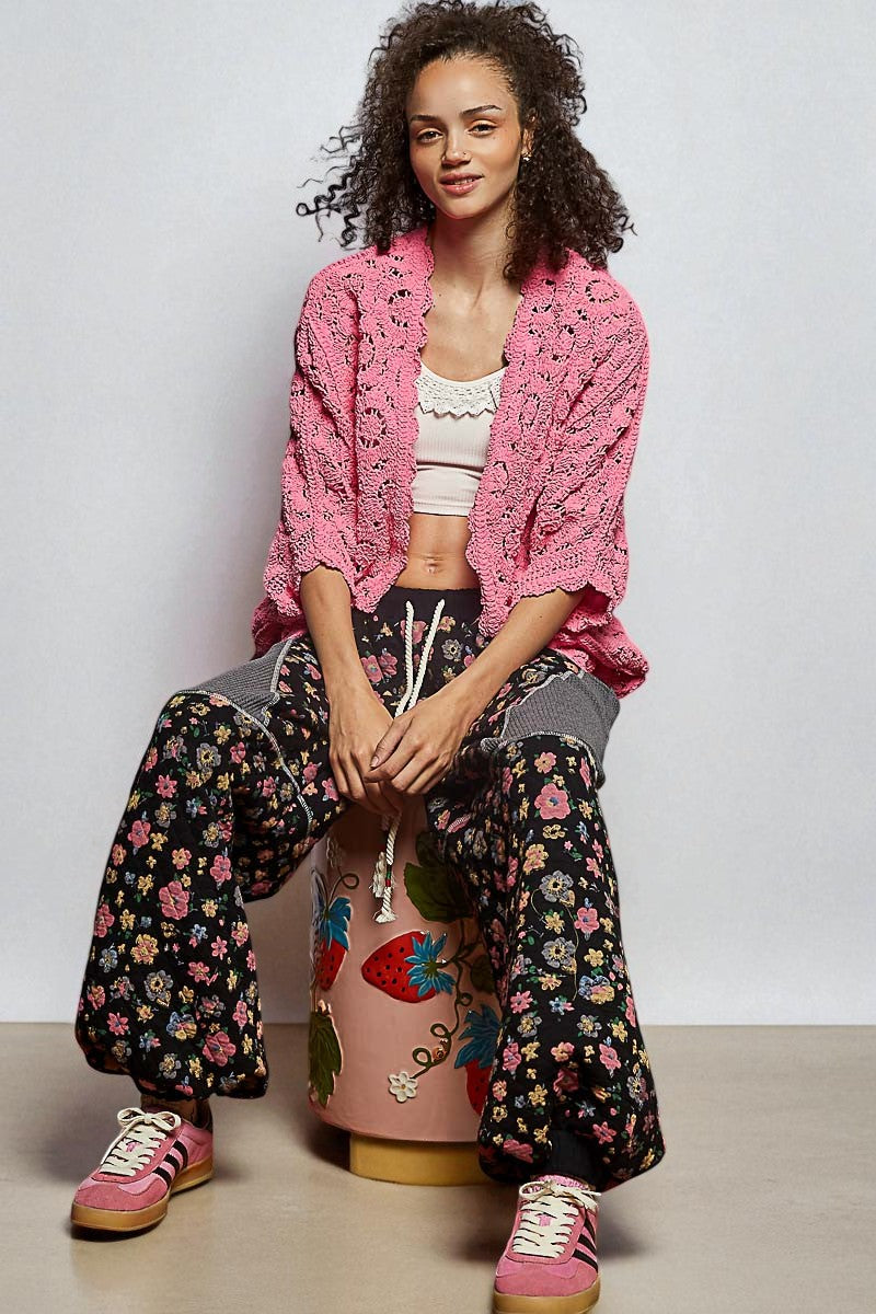 Woman sitting on a colorful stool wearing a pink crochet cardigan, floral pants, and pink sneakers.