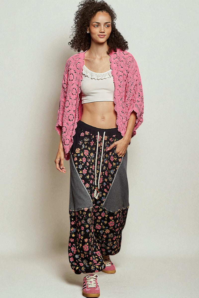 Woman wearing a pink crochet cardigan, white top, and floral pants on a plain background