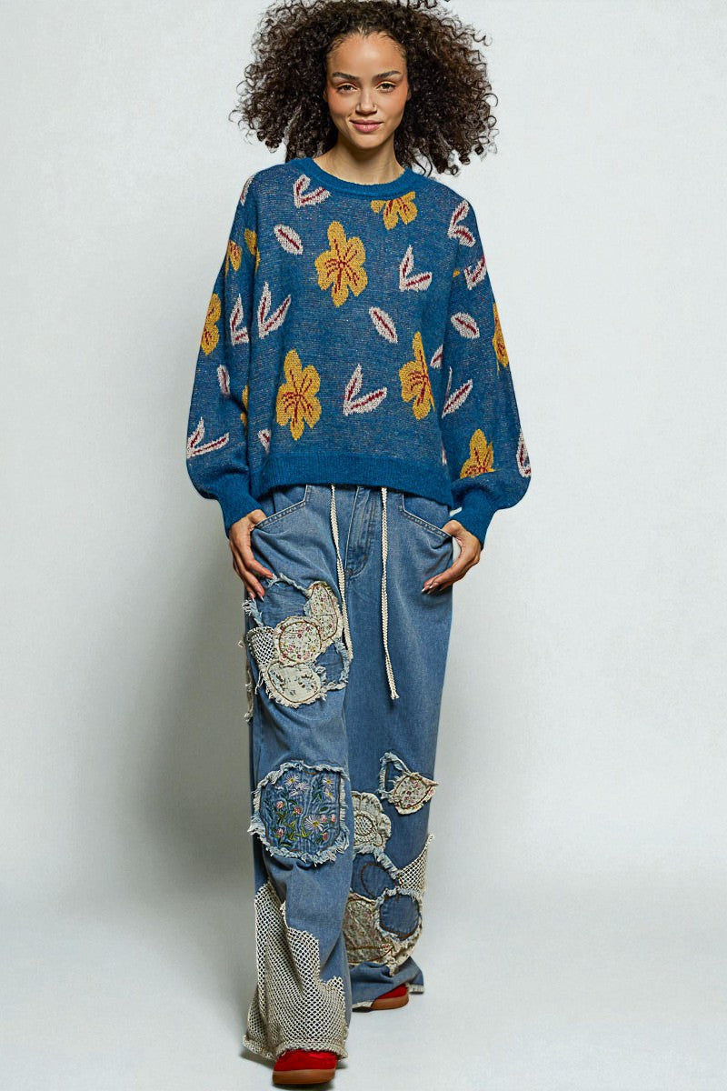 Person wearing a blue sweater with floral patterns and patchwork jeans on a white background