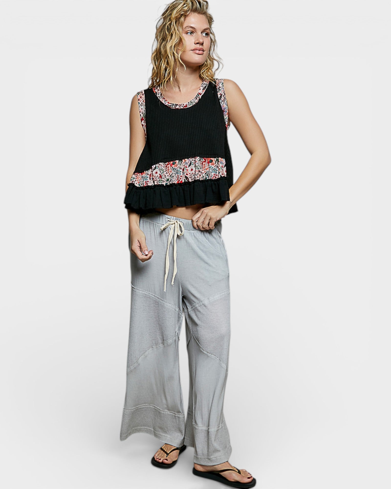 Woman wearing a black sleeveless top with floral patterns and gray wide-leg pants on a white background