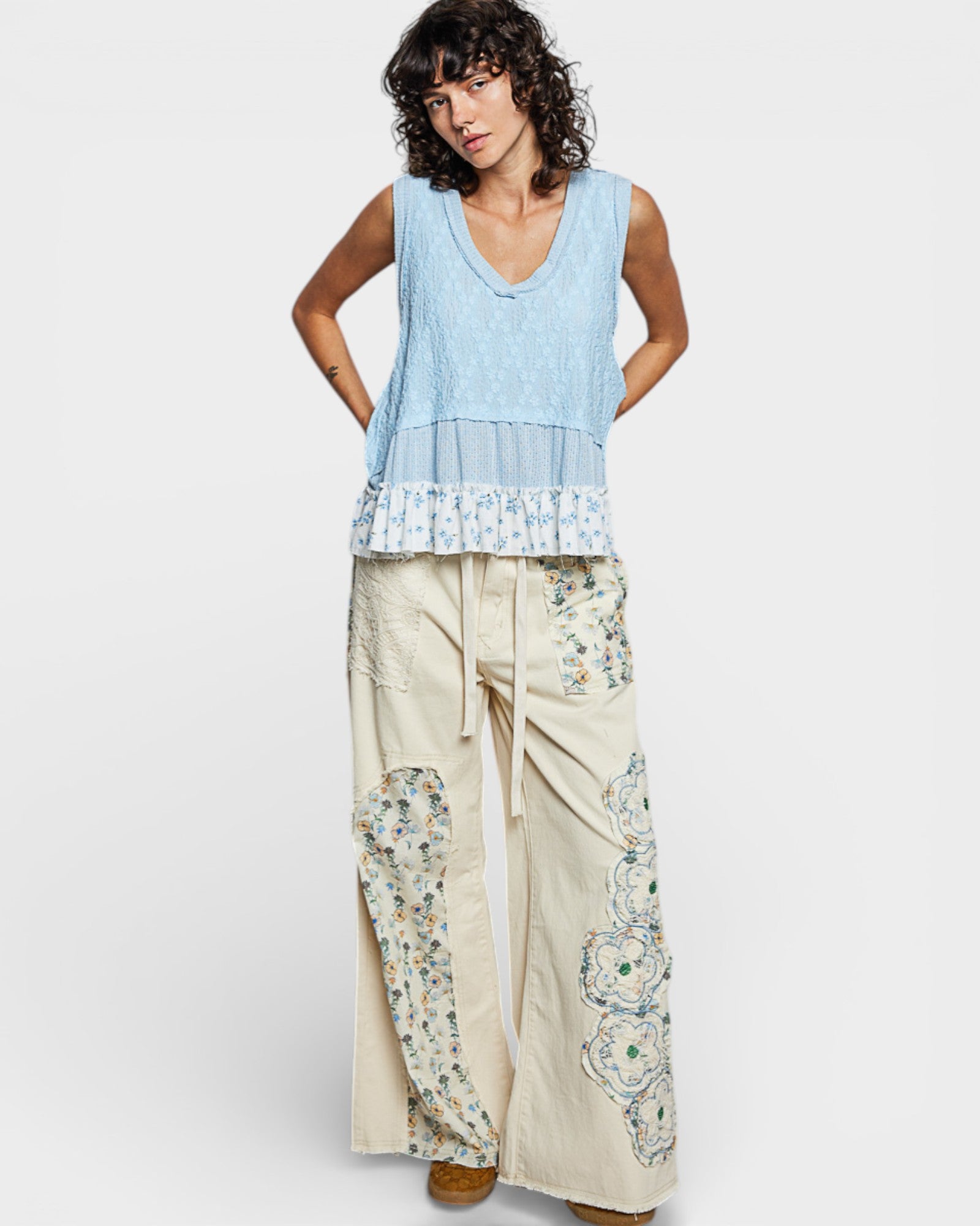 Woman wearing a light blue sleeveless top and beige pants with floral patterns on a white background