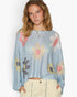 A woman wearing a light blue knit long-sleeve crop top with a colorful floral pattern in pink, yellow, and gray, paired with cream-colored pants, against a plain white background.