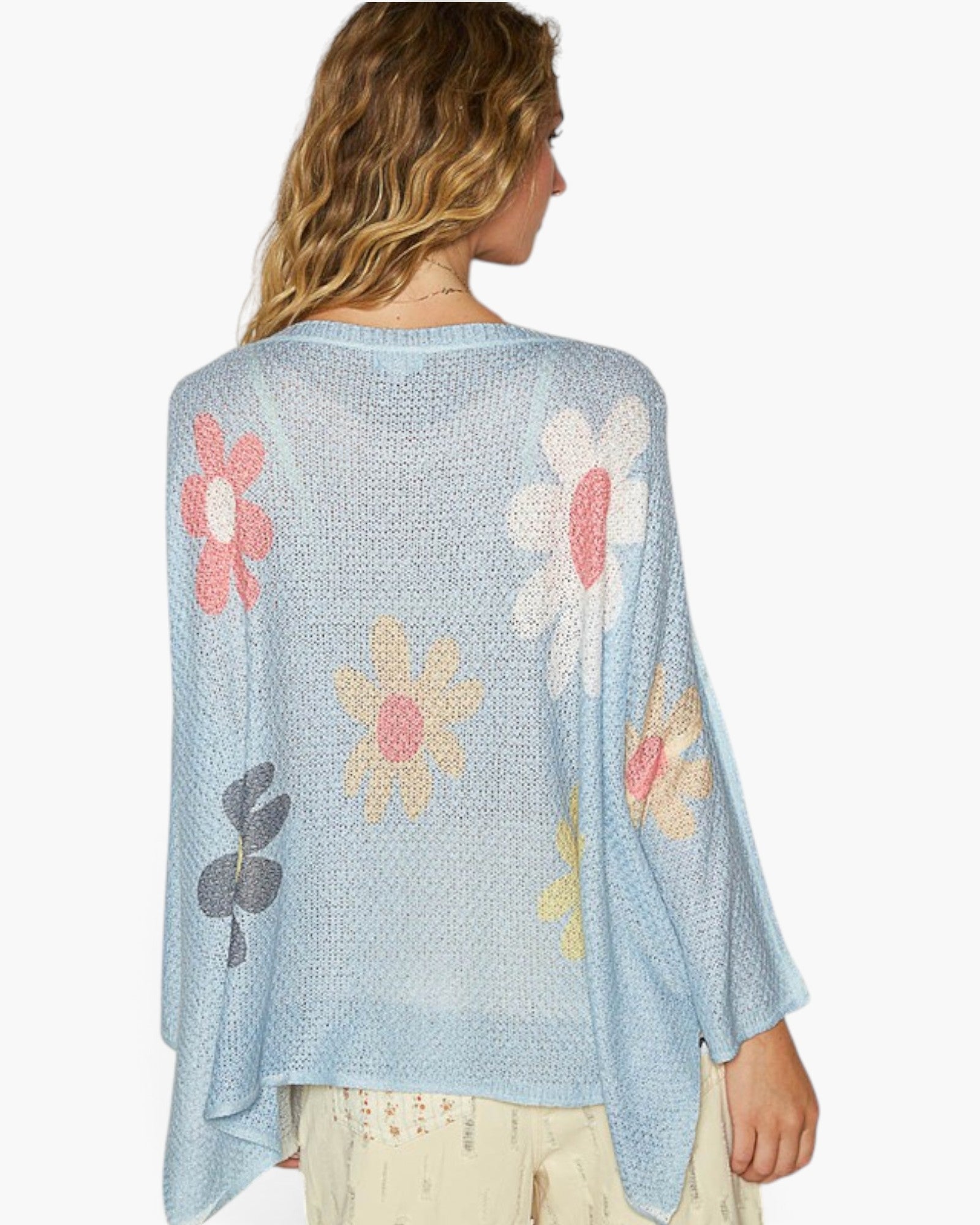 A woman shown from the back wearing a light blue open-knit poncho-style cardigan with large floral motifs in pink, yellow, white, and gray, paired with light beige pants against a plain white background.
