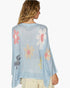 A woman shown from the back wearing a light blue open-knit poncho-style cardigan with large floral motifs in pink, yellow, white, and gray, paired with light beige pants against a plain white background.