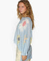 A woman wearing a light blue knit poncho sweater with a large daisy and sunflower pattern, paired with cream pants, photographed against a plain white studio background.
