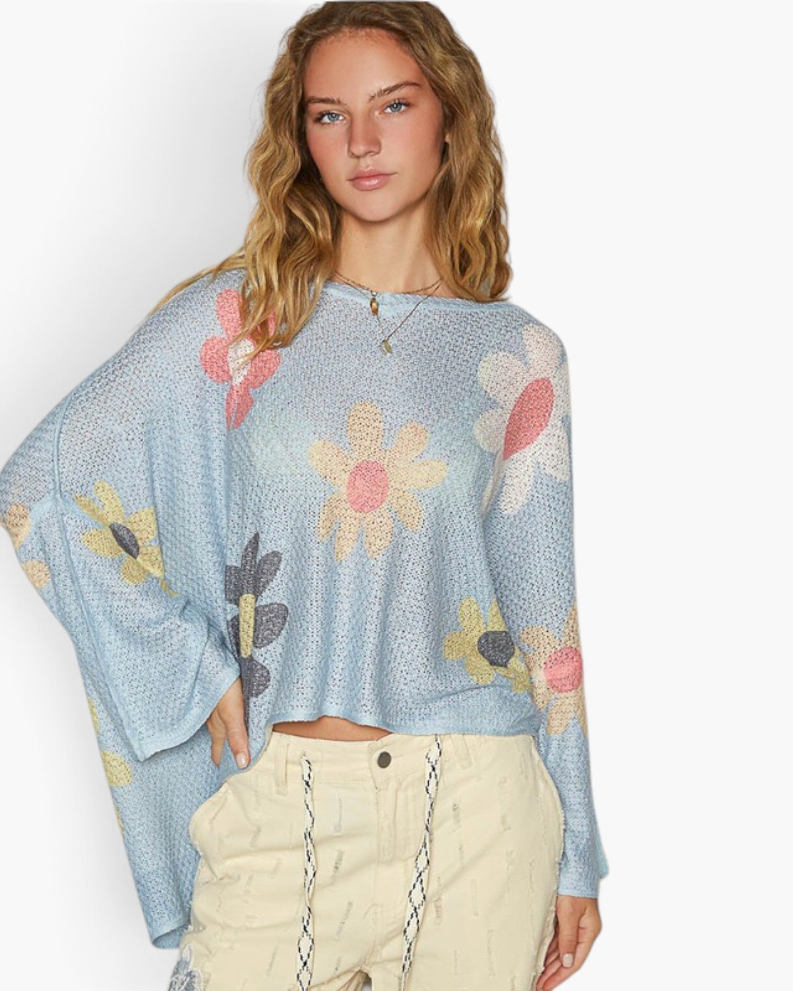 A woman modeling a light blue open-knit long-sleeve crop top with a colorful floral pattern, paired with cream-colored pants, against a plain white studio background.