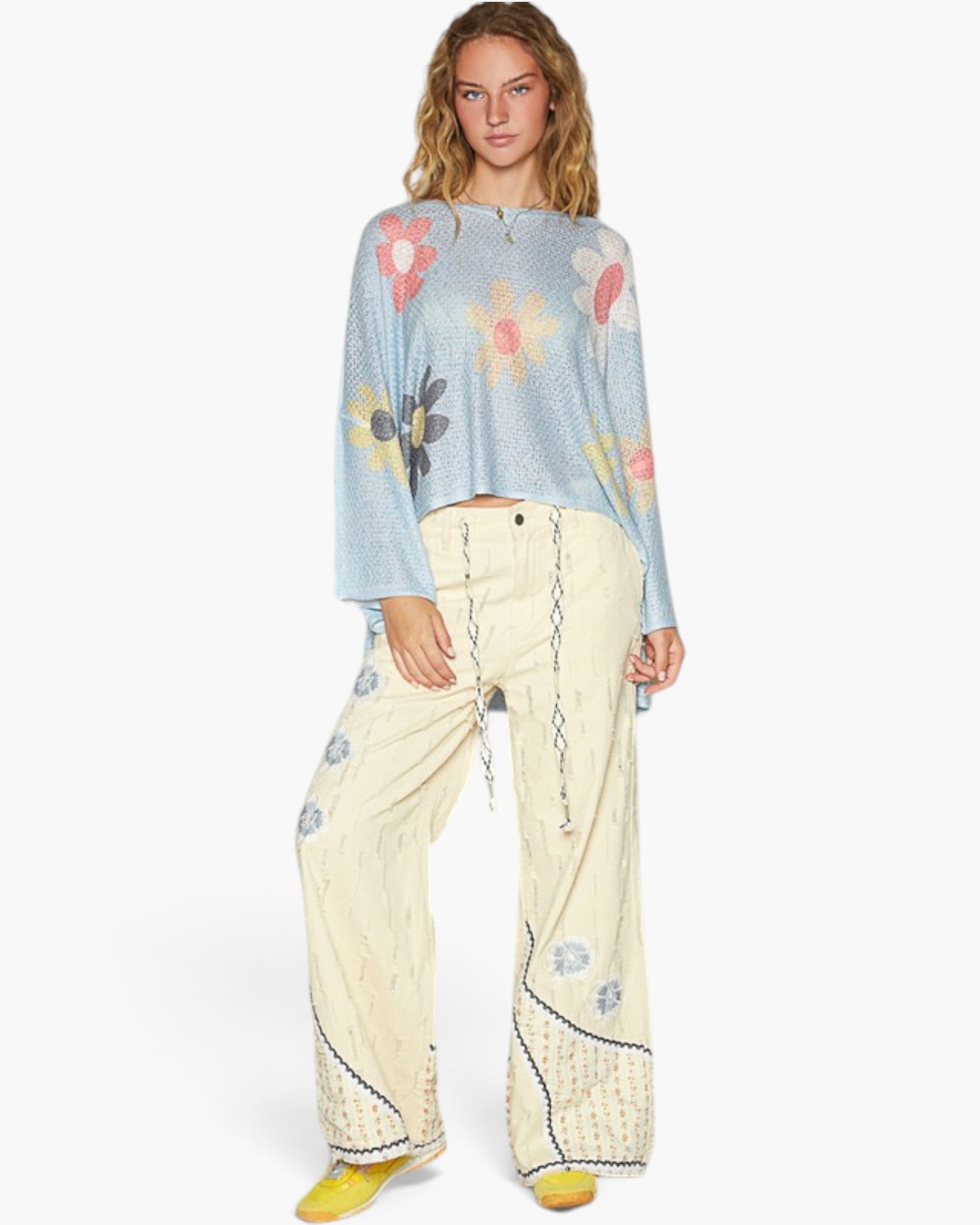 A model wearing a light blue long-sleeve knit top with a colorful floral pattern, paired with cream wide-leg pants featuring blue floral embroidery and drawstrings, and yellow shoes, against a plain white background.