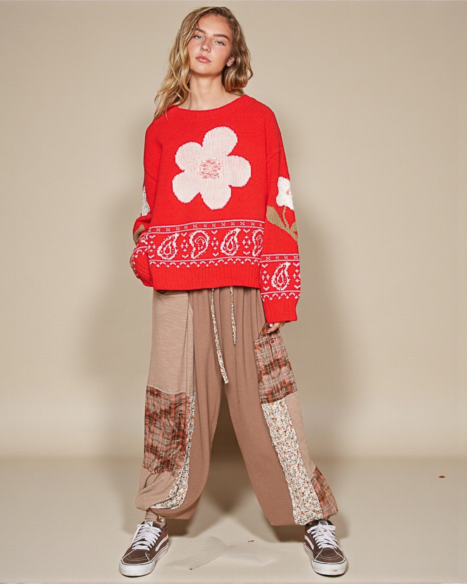 Person wearing a red sweater with a white flower design and patterned pants on a beige background