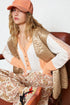 Woman wearing a beige quilted jacket, white shirt, and floral pants sitting on a brown chair.