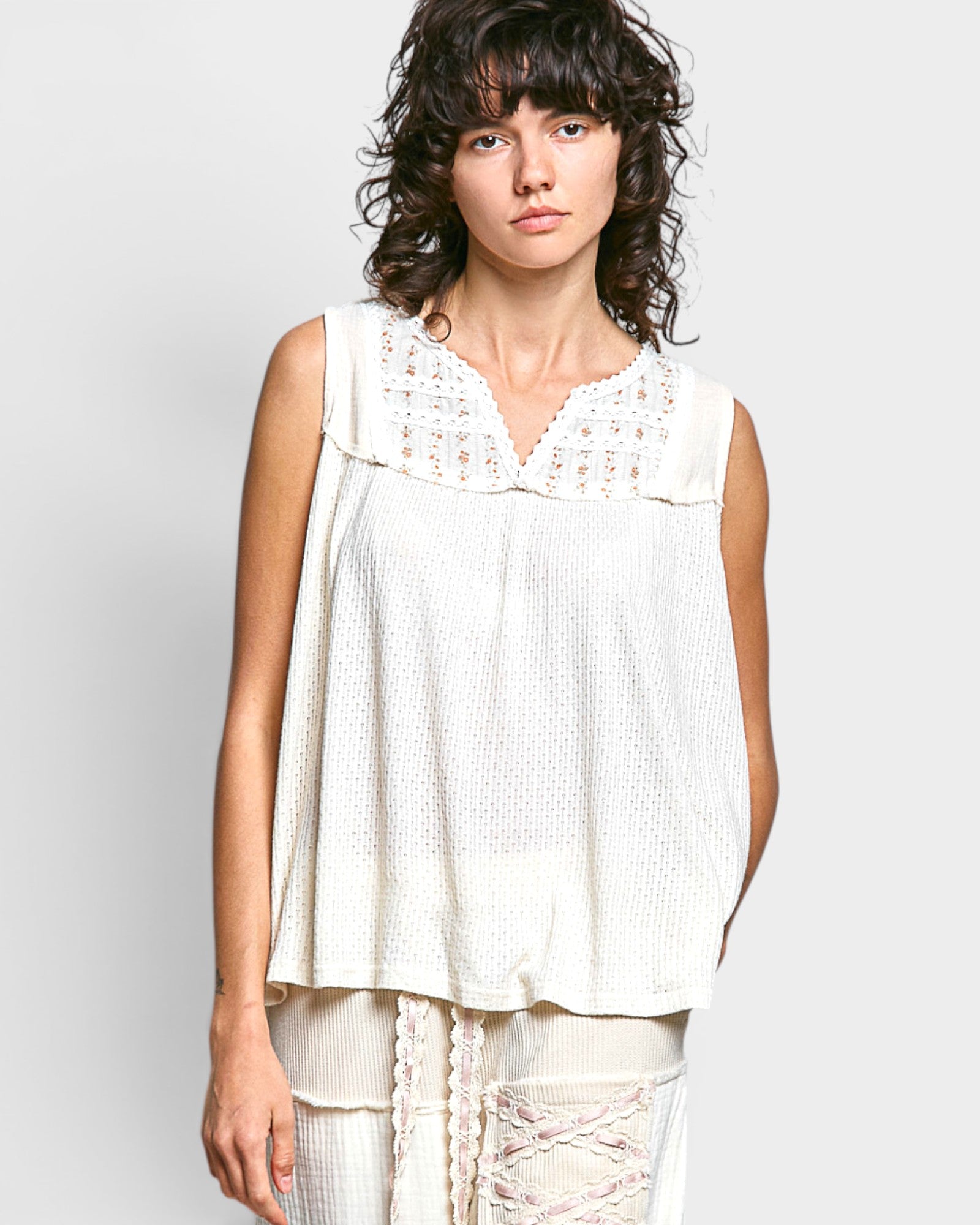 Woman wearing a white sleeveless top with lace details on a plain background