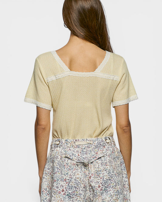 Woman wearing a light yellow textured top with lace trim and floral pants on a white background