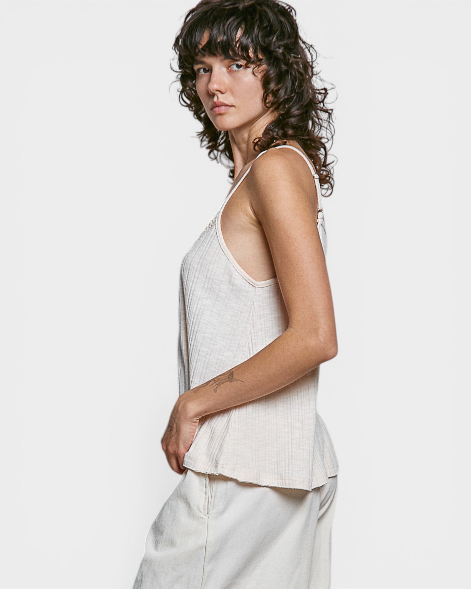 Woman wearing a light-colored sleeveless top and white pants against a plain background