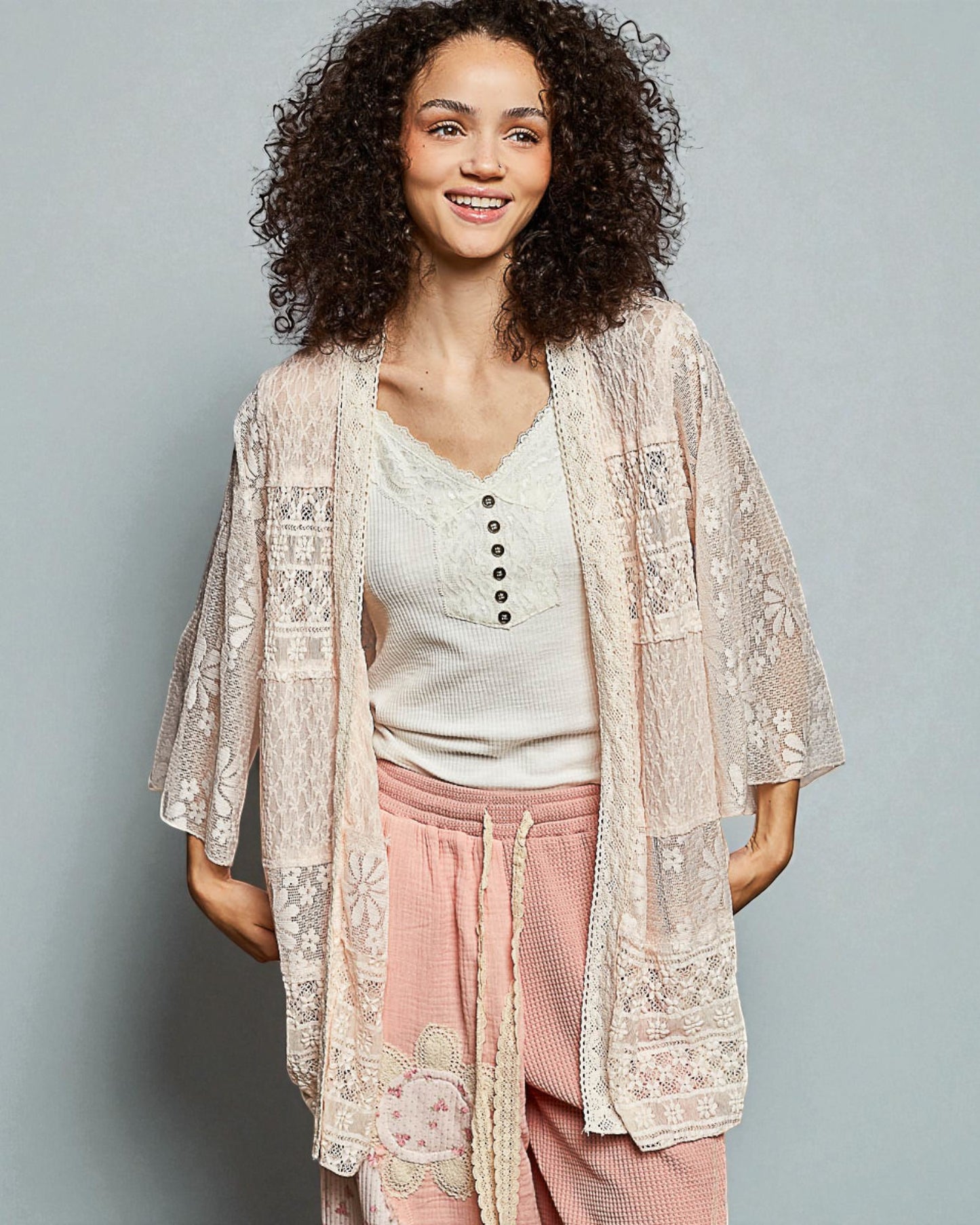 Woman wearing a lace kimono over a white top and pink pants on a gray background