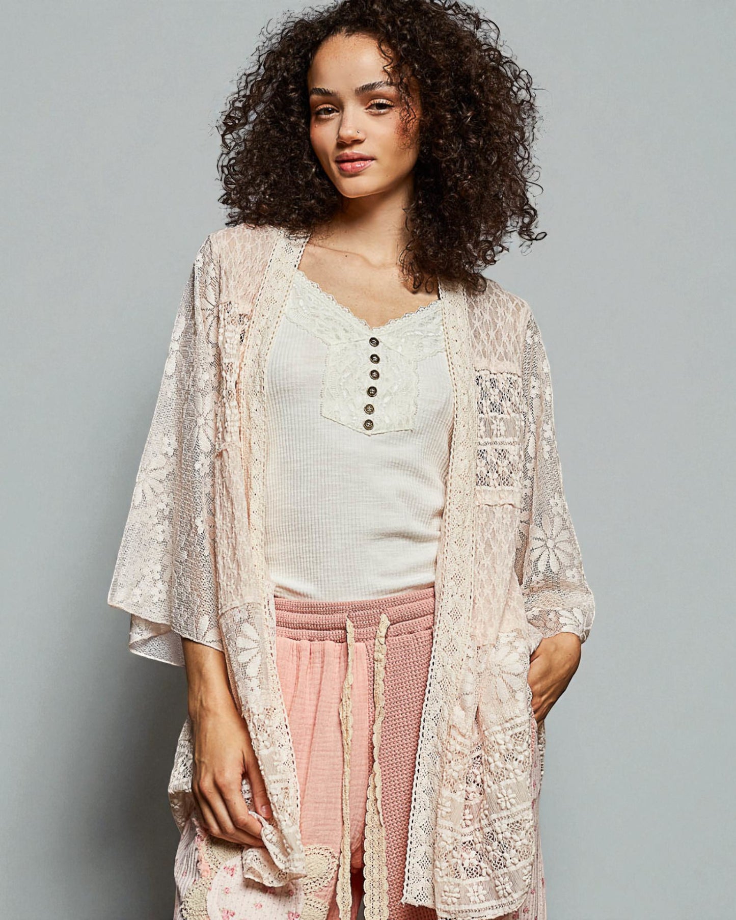 Woman wearing a lace kimono over a white top and pink pants on a gray background