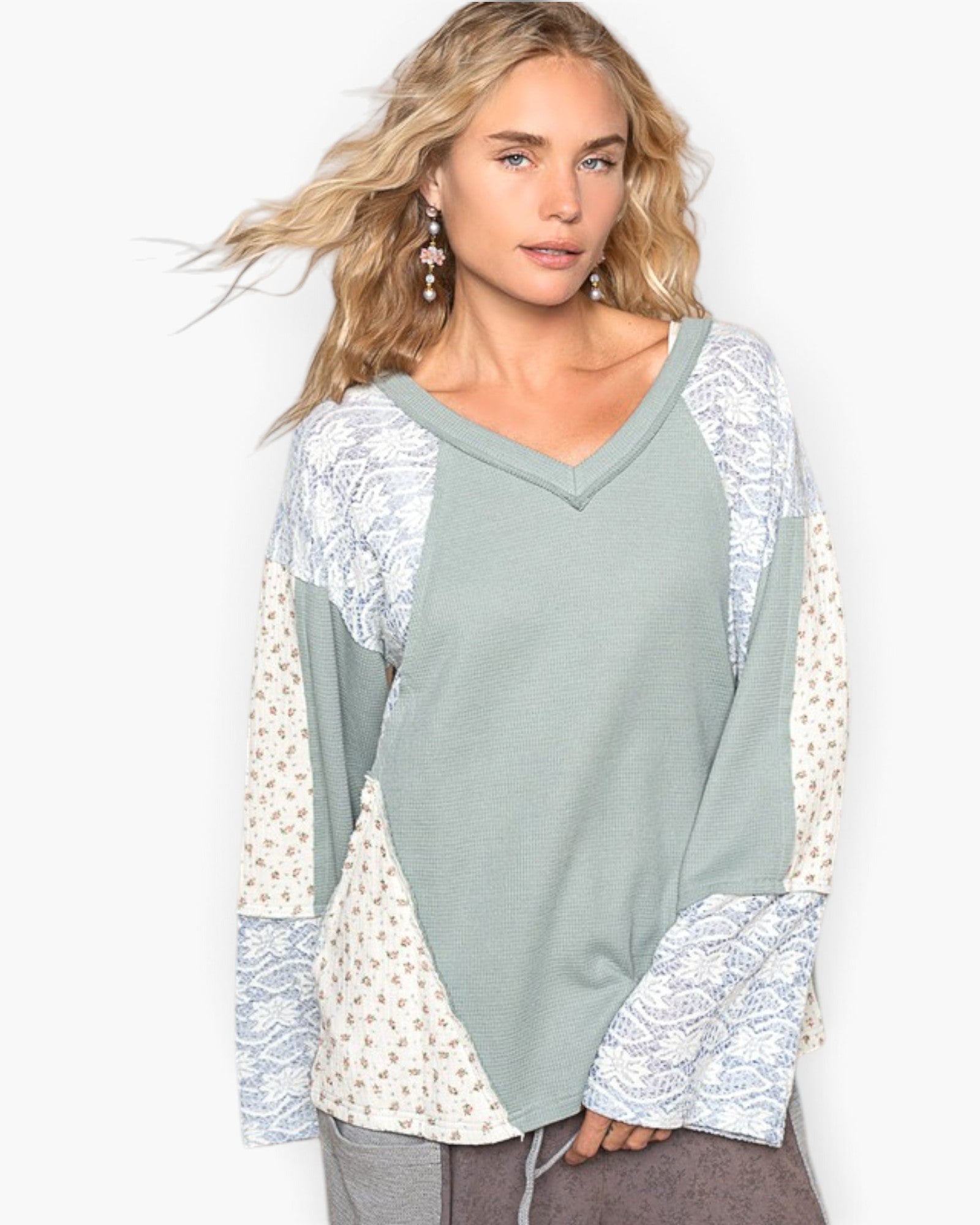 A woman modeling a long-sleeve V-neck top with a sage green body and cream-colored patterned sleeves and side panels, set against a plain white background.