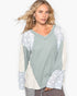 A woman modeling a long-sleeve V-neck top with a sage green body and cream-colored patterned sleeves and side panels, set against a plain white background.