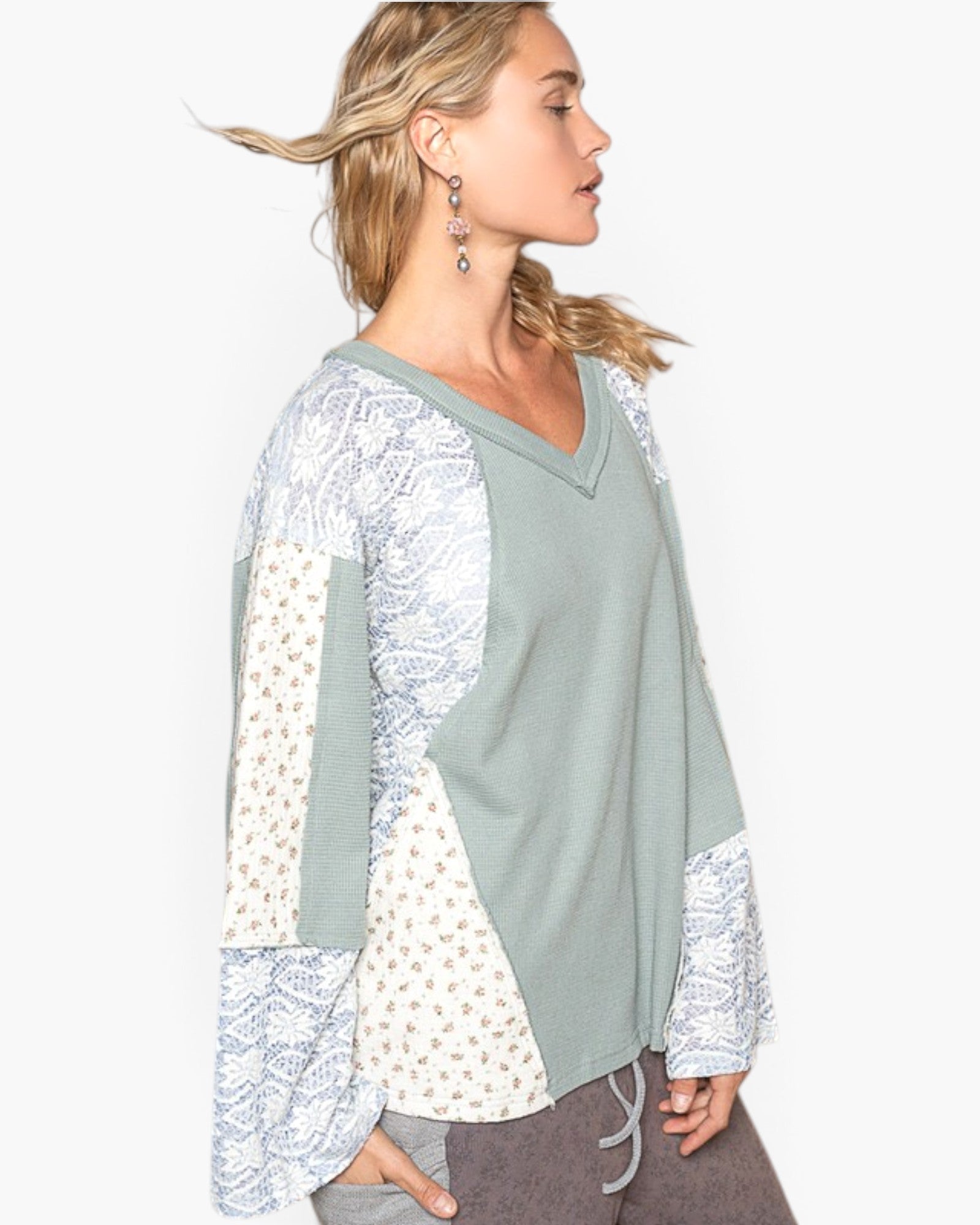 A woman shown in profile wearing a long-sleeve V-neck knit top with sage green body panels and mixed patterned sleeves and side panels in cream and light blue-gray.