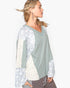 A woman shown in profile wearing a long-sleeve V-neck knit top with sage green body panels and mixed patterned sleeves and side panels in cream and light blue-gray.