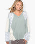 A woman wearing a sage green long-sleeve top with cream and blue patterned raglan sleeves, paired with gray drawstring pants, against a plain white background.