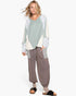 A woman modeling a patchwork long-sleeve top with sage green, cream, and floral patterns, paired with brown harem-style jogger pants and black slide sandals, against a plain white background.