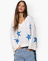 A woman wearing a cream-colored knit sweater with blue and pink embroidered starfish patterns, paired with light blue jeans, against a plain white background.