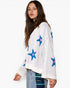 A woman wearing a white knit sweater with blue and pink star patterns, paired with light blue jeans, photographed against a plain white studio background.
