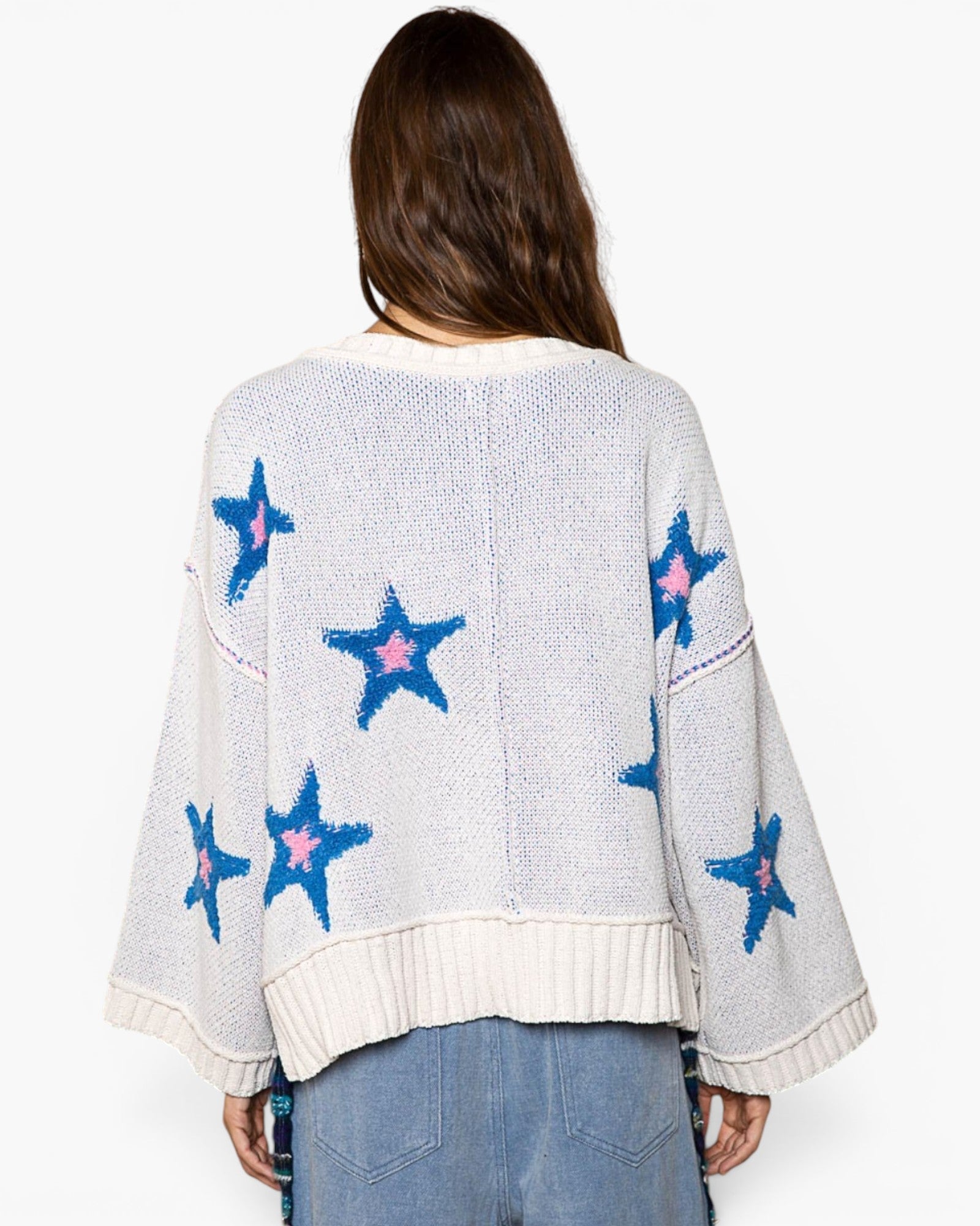 A woman viewed from the back wearing a cream knit sweater with blue and pink star motifs, paired with light blue jeans against a plain white background.