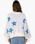 A woman viewed from the back wearing a cream knit sweater with blue and pink star motifs, paired with light blue jeans against a plain white background.