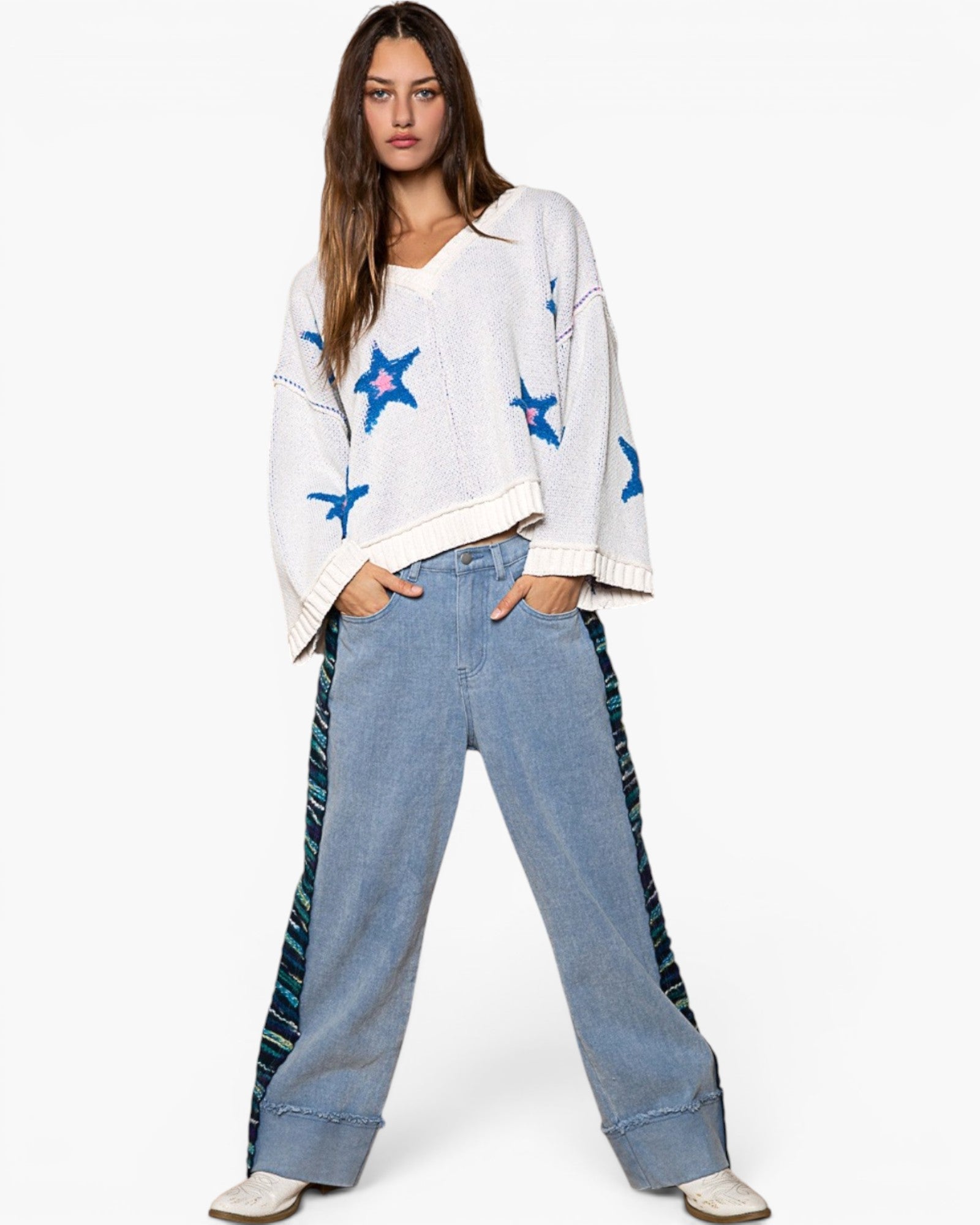 A woman modeling a white knit sweater with blue embroidered star patterns, paired with wide-leg light blue jeans featuring dark patterned side panels, against a plain white studio background.