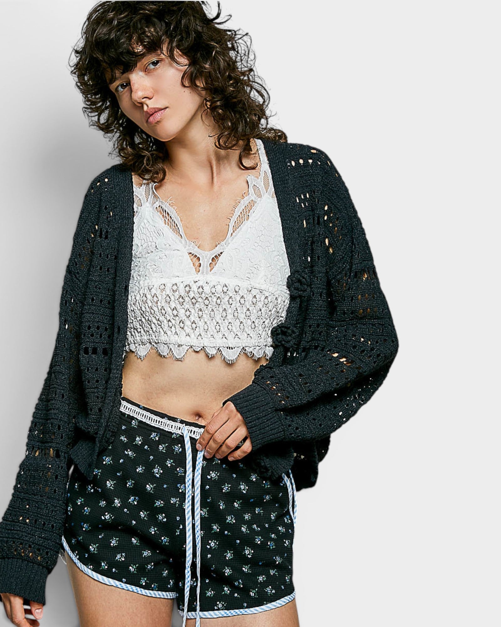 Woman wearing a black crochet cardigan, white lace top, and patterned shorts on a light gray background