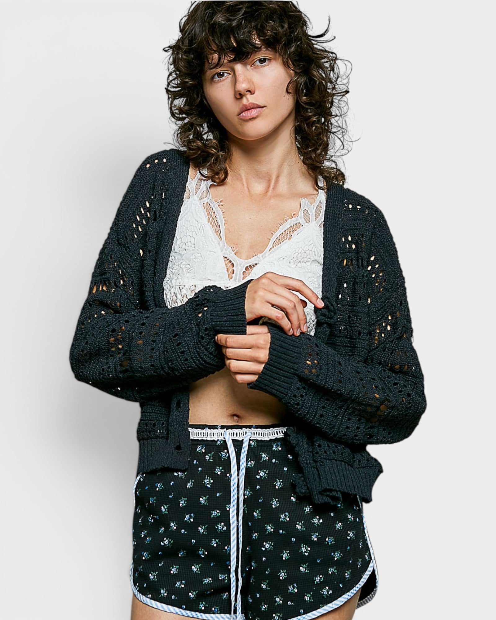 Woman wearing a dark green cardigan over a white lace top and patterned shorts on a light gray background