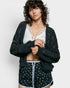 Woman wearing a dark green cardigan over a white lace top and patterned shorts on a light gray background