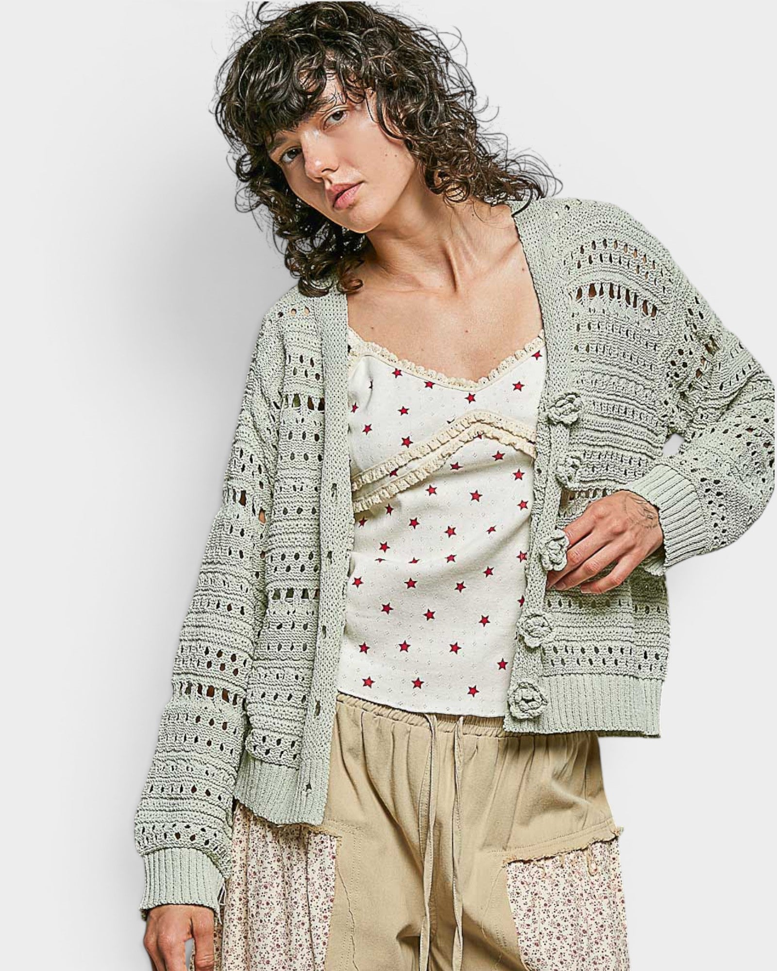 Woman wearing a light green crochet cardigan over a white top with red patterns and beige pants on a plain background.