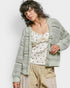 Woman wearing a light green crochet cardigan over a white top with red patterns and beige pants on a plain background.