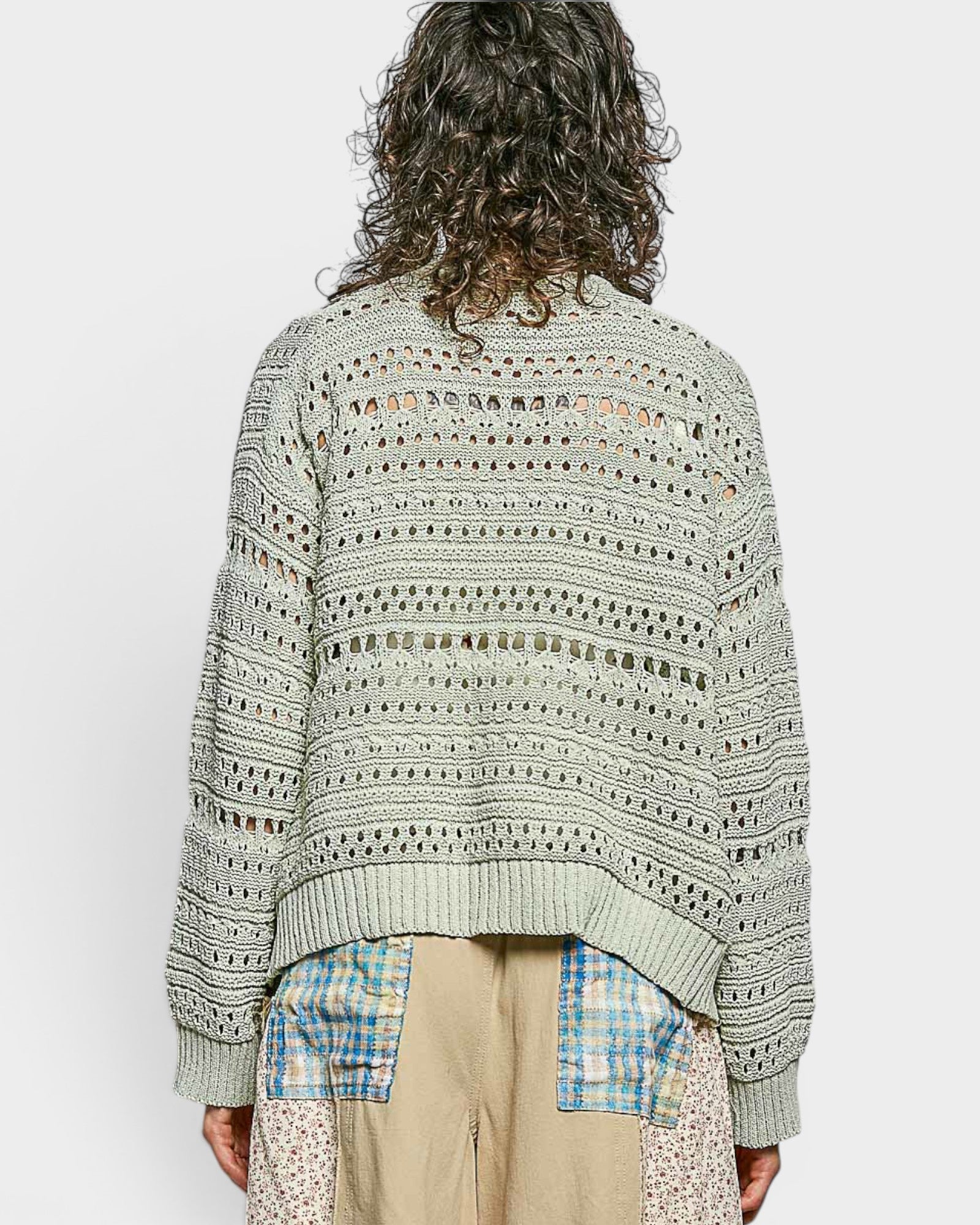 Person wearing a patterned sweater over a plaid shirt and beige pants on a white background