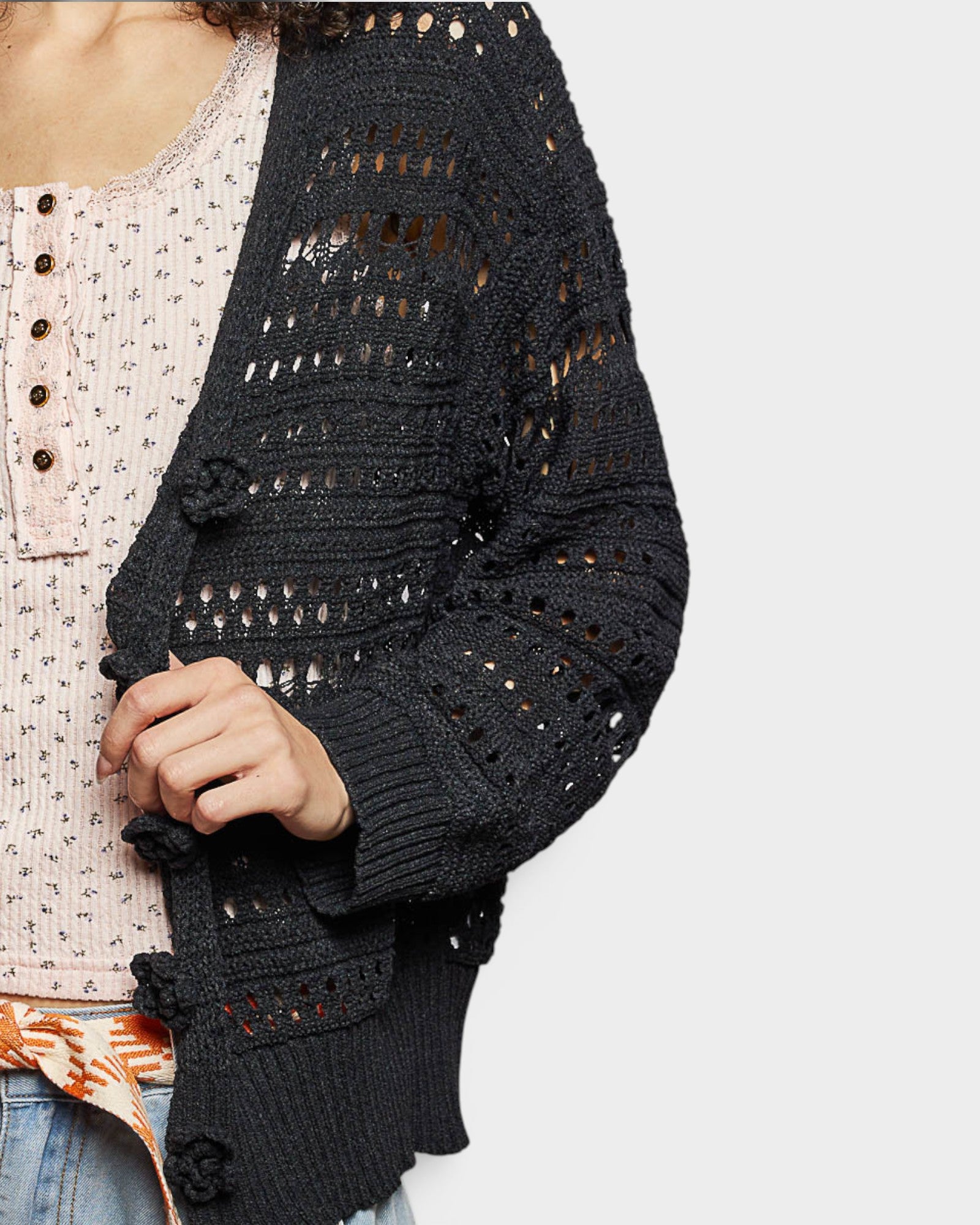 Black open-knit cardigan worn over a patterned top on a light gray background