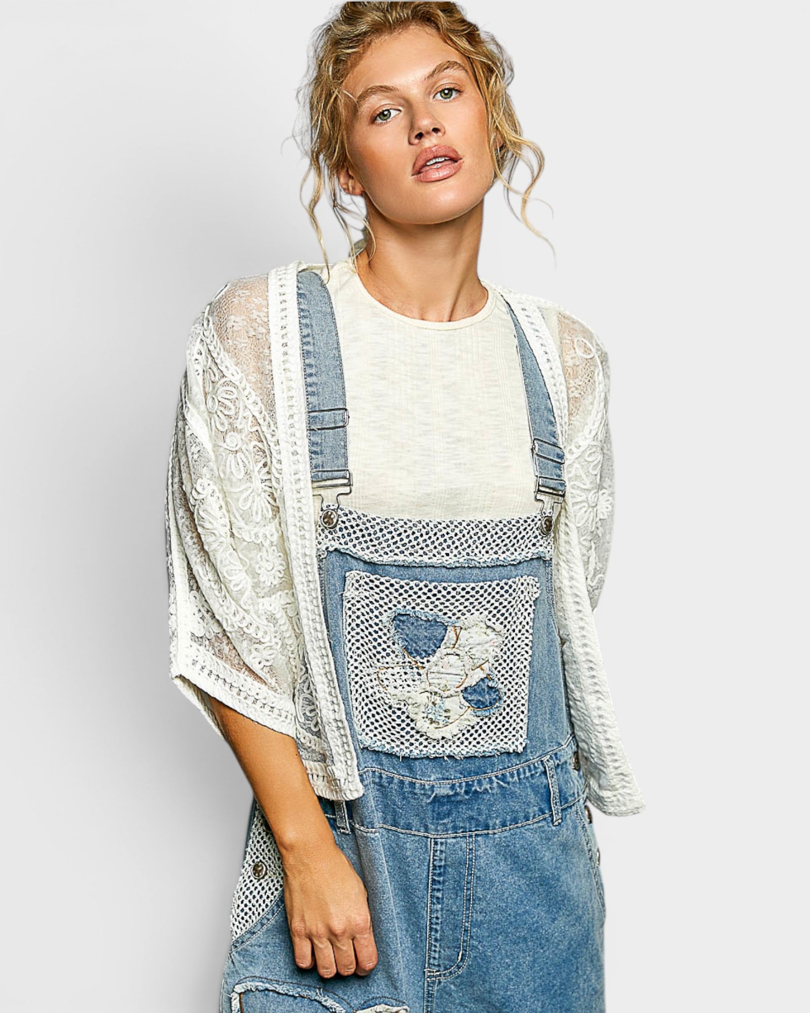 Woman wearing a white lace top and blue denim overalls with floral embroidery on a light gray background