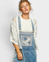 Woman wearing a white lace top and blue denim overalls with floral embroidery on a light gray background